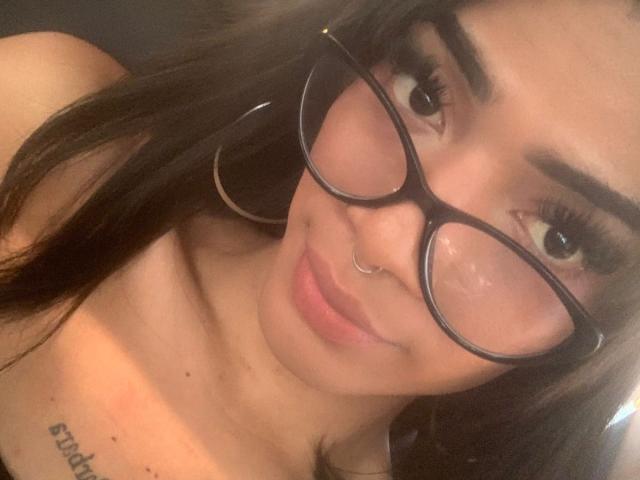 DeboraRaquel, usando óculos, com um piercing no nariz e brincos de argola, sorrindo para a câmera, mostrando seus seios e uma pequena tatuagem no lado direito. Olhos castanhos, cílios longos..