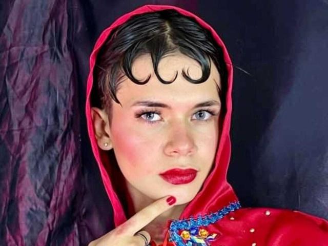 JazmiinRosee with short black curly hair, blue eyes, red lipstick, red hood, and red dress with blue embroidery, pointing at her chin with a finger.