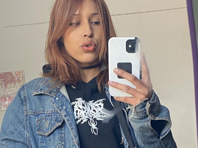 KiaraHernandezX mit braunen Haaren, voller Lippen, einer schwarzen Halskette, einer Jeansjacke und einem schwarzen T-Shirt mit Aufdruck, hält ein weißes Telefon und macht ein Selfie. Ihr Hintern ist nicht sichtbar..