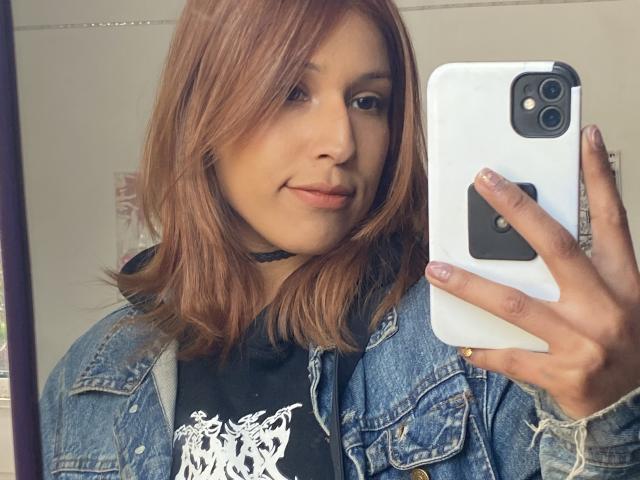 KiaraHernandezX, com cabelo castanho, vestindo uma jaqueta jeans sobre uma camisa preta, tirando uma selfie com um celular branco, tem um sorriso discreto e um colar preto..