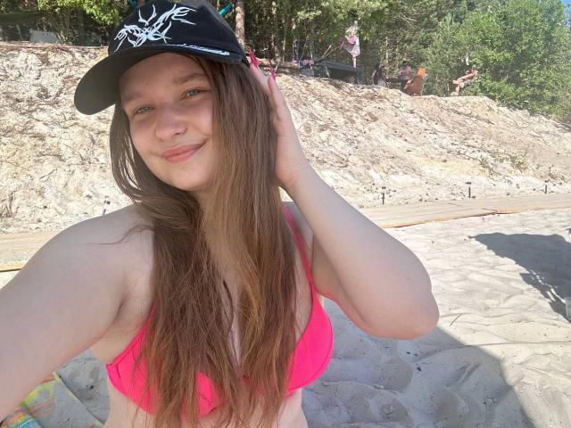 MagikPeachh mit langen braunen Haaren, lächelnd, trägt einen schwarzen Hut mit weißem Muster, ein rosa Bikini-Oberteil, zeigt kleine Brüste, steht auf einem sandigen Strand und hält den Hut..