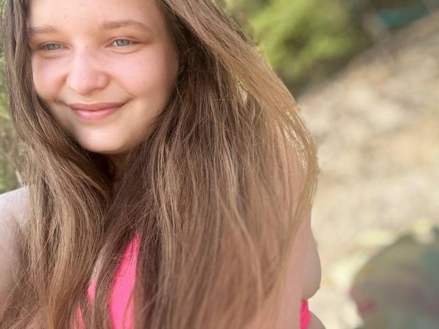 MagikPeachh hat langes, braunes Haar, blaue Augen und ein leichtes Lächeln. Sie trägt ein pinkes Oberteil, bei dem ihre Brüste teilweise sichtbar sind, und befindet sich in einer sonnigen, offenen Umgebung..