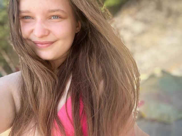 MagikPeachh mit langen braunen Haaren, lächelnd, mit blauen Augen, trägt ein pinkes Oberteil, wobei ihre Brüste leicht zu sehen sind, draußen im Sonnenlicht..