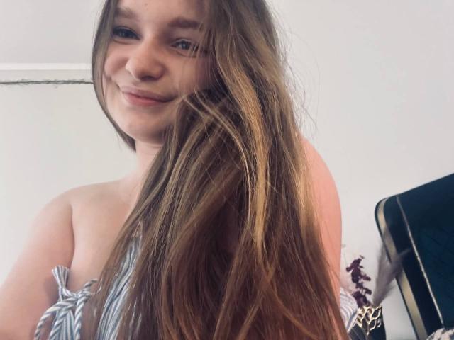 MagikPeachh con lunghi capelli castani, sorridente, che indossa una maglietta a righe, mostra piccoli seni, pelle chiara e occhi azzurri, in un selfie casuale..