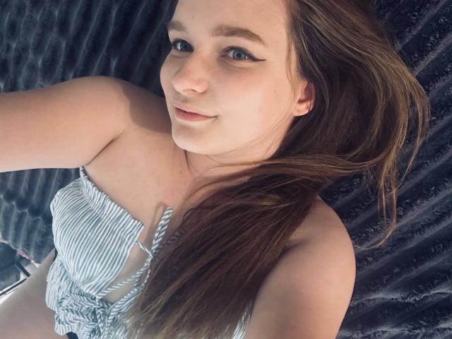 MagikPeachh mit langen braunen Haaren, blauen Augen und dezentem Eyeliner, trägt ein gestreiftes Oberteil, lächelt leicht und liegt auf einer dunklen, strukturierten Oberfläche, wobei ihre nackten Schultern zu sehen sind..