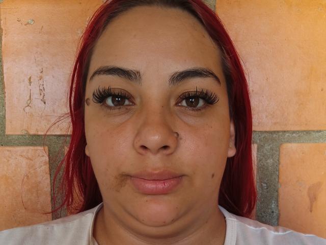LaGorditaPelirRoja tem cabelo ruivo, olhos castanhos, cílios longos, um piercing no nariz e uma pequena pinta na bochecha direita, vestindo uma camisa branca e olhando diretamente para a câmera..