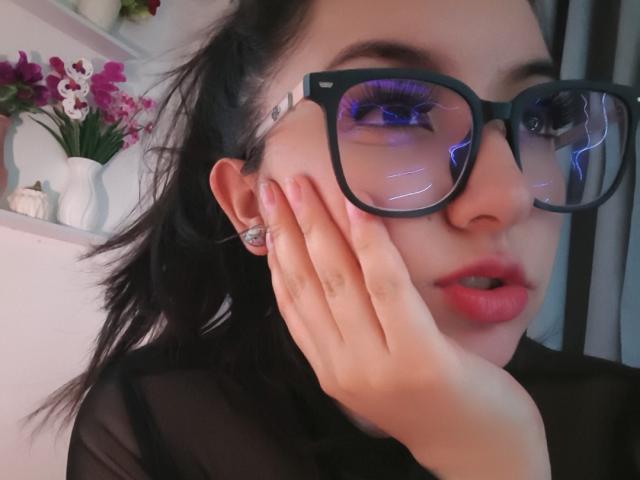 SusanMonroy with black glasses, pink lips, and dark hair, touching her cheek, showing a diamond earring, wearing a sheer black top, with purple reflection in glasses.