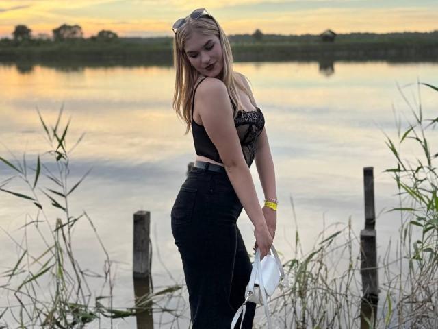 AlinaLemon está de pie junto a un canal, vestida con una blusa negra de encaje y jeans de cintura alta, sosteniendo una camisa blanca, con cabello rubio, una pulsera amarilla y gafas de sol en la cabeza..