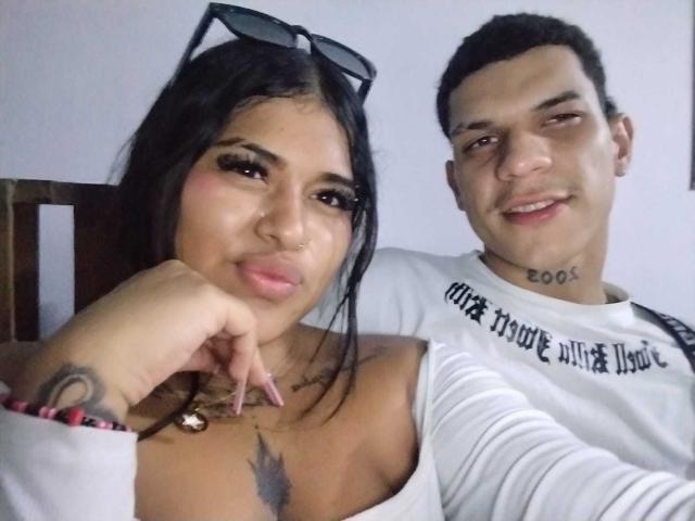 PartyLindaDestruction, con los senos a la vista en una blusa blanca, cabello negro, gafas en la cabeza, un tatuaje en la muñeca y un collar. El novio de PartyLindaDestruction, cabello negro corto, camisa blanca, tatuaje en el cuello..