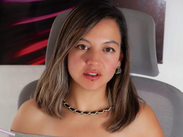 ArianaBlonde hat schulterlanges, braunes Haar, eine Kette um den Hals und eine Lippenpiercing, mit kleinen Brüsten und braunen Augen, und sitzt auf einem grauen Bürostuhl..