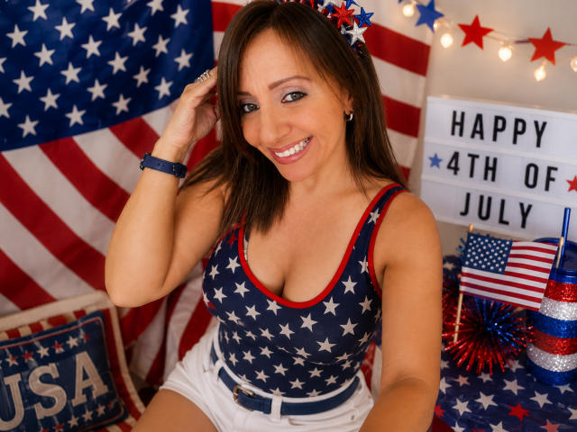 Charlotte Dilucca, con i capelli castani, sorridente, indossa una canottiera con stelle e strisce, mostra il seno, pantaloncini bianchi e una fascia rossa e blu tra i capelli, con uno sfondo a stelle e strisce americane..