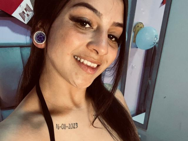 LonxEva with dark brown hair, wearing black halter top, smiling, blue circular ear gauges, small tattoo below neck, slightly parted lips, and visible skin texture.