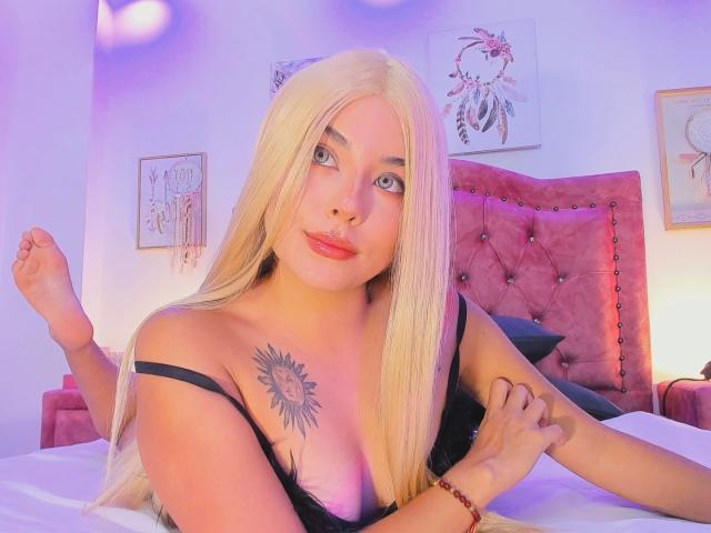 GraceClarke with long blonde hair, black lingerie, sun tattoo on chest, pink headboard, and red bracelet, laying on bed, looking up, right arm raised, left arm on bed.