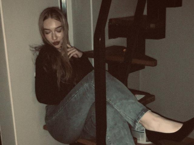 JoetteShigeta sits on wooden stairs, legs crossed, wearing a black long-sleeve top and blue jeans, black socks, with long blonde hair and dark lipstick, looking down.