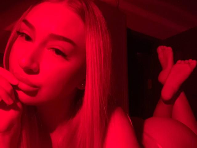 Red-lit photo of JoetteShigeta with blonde hair, lying down, legs up, fingers near mouth, soft smile, bare shoulders, feet in air, relaxed, sexy, confident, subtle eyeliner.