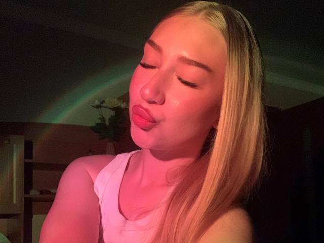 JoetteShigeta with blonde hair, pinkish-red skin, closed eyes, puckered lips, wearing a white tank top, illuminated by a green-red light, in a dimly lit room.