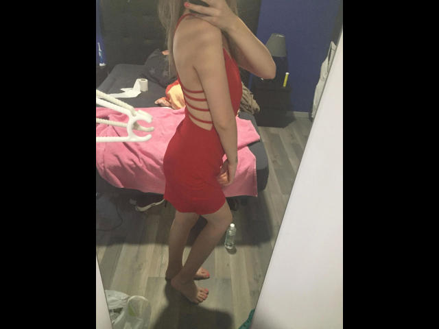 Studentessa, molto attraente in un vestito rosso con spacchi laterali, in piedi a piedi nudi in un selfie allo specchio, che mostra il sedere e piccoli seni, in una camera da letto con un letto rosa e una bottiglia..