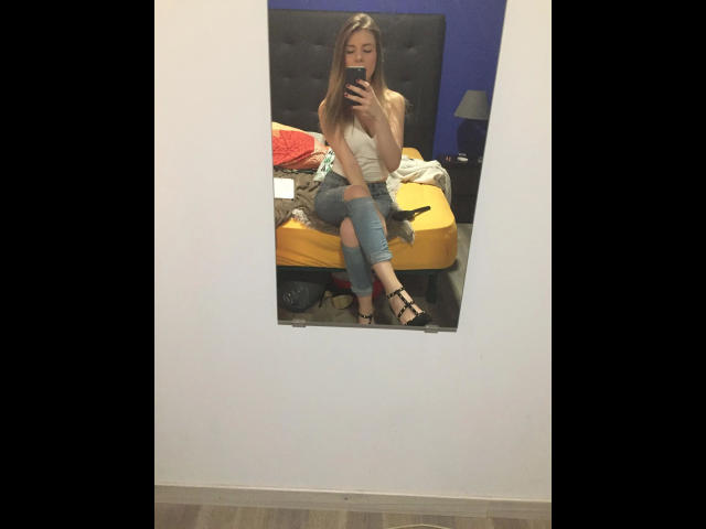 Studentin - Eine junge Frau mit langen, braunen Haaren, kleinen Brüsten, die ein weißes Tanktop, blaue Jeans und schwarze High Heels trägt und ein Selfie vor einem Spiegel in einem unordentlichen Schlafzimmer macht..