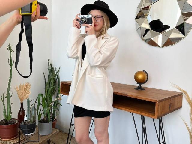 Felicita Zakrajsek, con capelli biondi, cappello nero, occhiali, giacca bianca, gonna nera, che tiene una macchina fotografica, piante in vaso, tavolo di legno, specchio, braccio di supporto per la macchina fotografica nell'inquadratura, tracolla gialla..