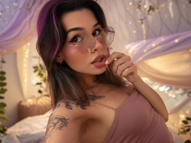 LenaBranch with pink-tinted hair, round glasses, and pink top, biting her finger, showing tattoos on her shoulder and chest, looking seductive. Boobs visible. Soft, warm lighting.