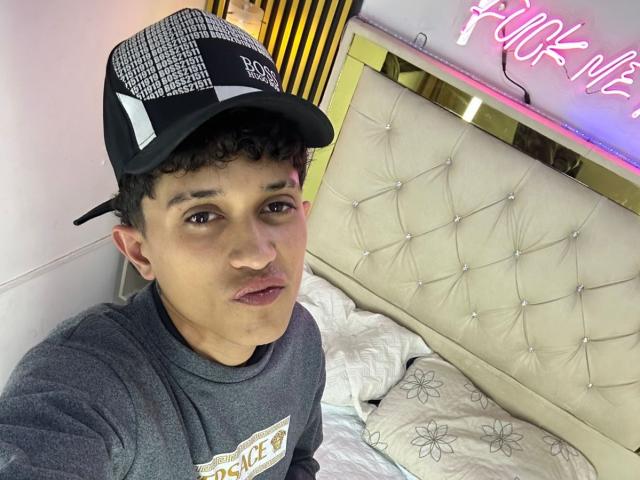 JaySecret, con una gorra de béisbol negra y blanca, una sudadera gris y cabello rizado, con una expresión neutra, está sentada en una cama beige con capitonado. En el fondo, hay un letrero de neón rosa..
