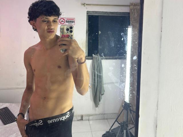 JaySecret, sin camisa, con cabello oscuro y rizado, sosteniendo un teléfono, vistiendo ropa interior negra con "emque2" en la cintura, selfie frente al espejo, el espejo refleja la habitación, paredes blancas, teclado negro a la izquierda..