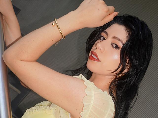 LindaMancini with black hair, red lipstick, and a gold bracelet, holding a pole, wearing a yellow, ruffled top, looking over her shoulder, showing her arm and shoulder.