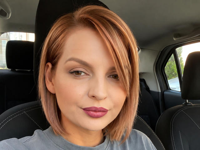 DouceAmelia ha capelli castano-rossicci di lunghezza media, pelle chiara e indossa un rossetto viola. È seduta in un'auto con sedili in pelle nera, e guarda leggermente di lato..