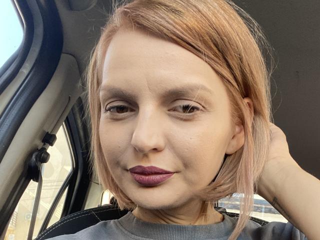 DouceAmelia has light brown, shoulder-length hair, wearing dark purple lipstick, and a gray shirt, with a hand in her hair, sitting in a car, looking slightly to the side.
