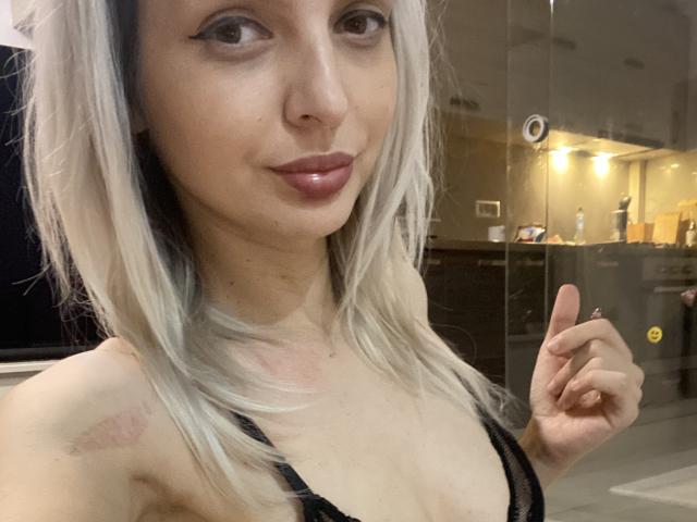 DouceAmelia, con i capelli biondo chiaro, un reggiseno di pizzo nero e un trucco leggero, sorride appena e indica con il dito indice, mostrando piccole cicatrici sul seno destro..