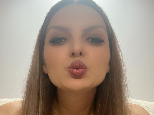 DouceAmelia, con labbra rosa carnose, occhi marroni e lunghi capelli castani, che fa una smorfia da bacio, con il naso leggermente sollevato, con un trucco molto leggero, su uno sfondo bianco uniforme..
