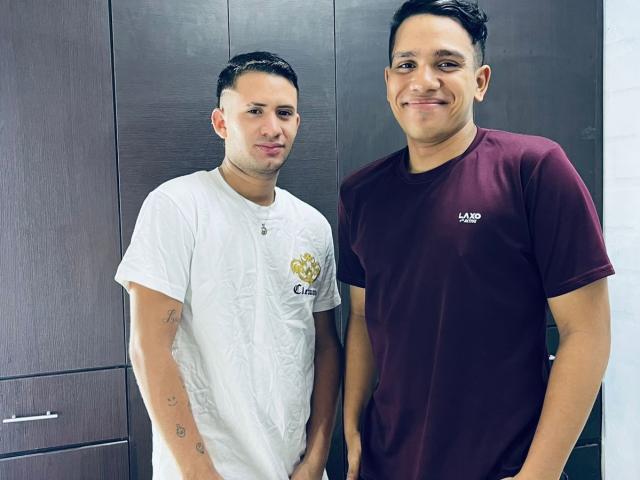 Due uomini, in piedi, che indossano magliette bianche e bordeaux, con capelli scuri corti, pelle bianca e marrone, tatuaggi sul braccio sinistro; la maglietta bordeaux dice "LAND"; entrambi sorridono, con uno sfondo di un armadio in legno..