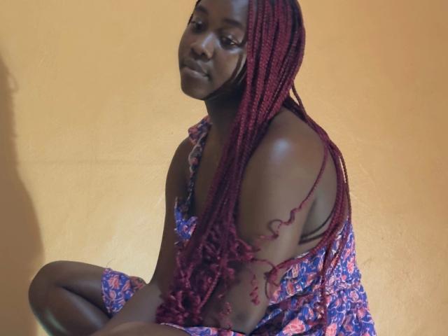 Sweetness26 - een aantrekkelijke vrouw met donkere huid en rode vlechten, zittend, gekleed in een blauwe jurk met bloemenprint, waarbij haar schouder en bovenarm onbedekt zijn, die naar beneden kijkt, voor een gele muur..