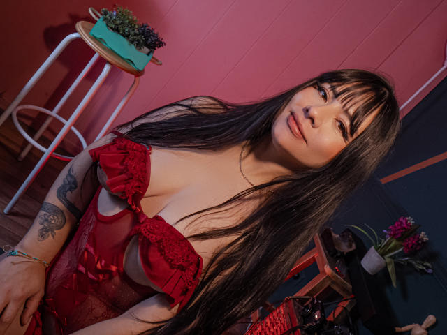 RoseAnna mit langen, schwarzen Haaren, die rote Spitzendessousards trägt und ihre Brüste zeigt, hat ein Tattoo an ihrem linken Arm, lächelt und befindet sich in einem rosafarbenen und blauen Raum mit einem Hocker..