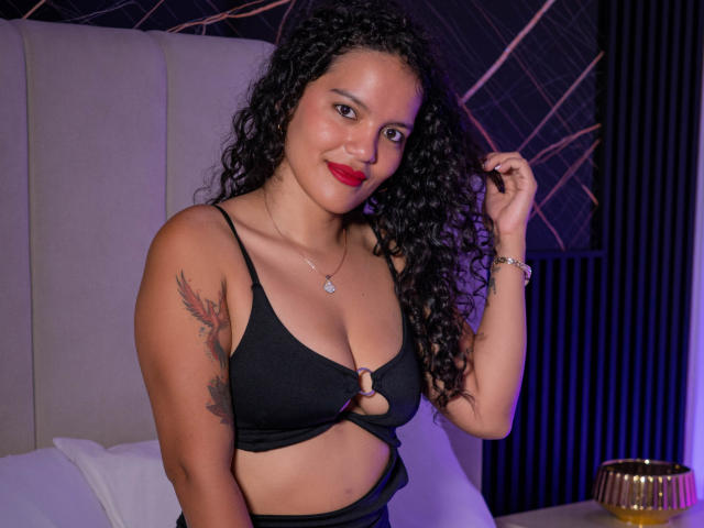 Amily Harlow mit lockigem, schwarzem Haar, rotem Lippenstift, schwarzer BH, Bügelcups, Tattoo am linken Arm, lächelnd, Haare haltend, Schlafzimmer, violette und goldene Beleuchtung, Schmuck, sexy..