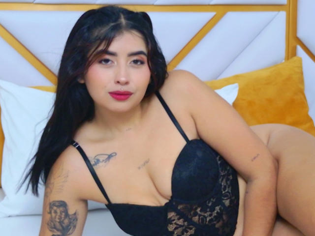 Valery Silva, con lencería de encaje negro, mostrando sus pechos, tatuajes en los brazos y las piernas, acostada en una cama con almohadas amarillas, mirando a la cámara con lápiz labial rojo..