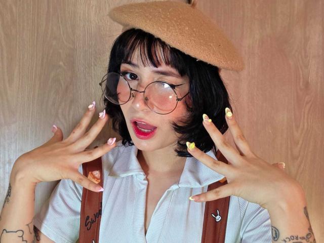 Miyuu69 mit schwarzem Bob, runder Brille, rotem Lippenstift, gelben Nägeln, weißem Hemd, brauner Beere, braunen Hosenträgern, Tattoos an den Händen, Mund offen, sieht süß aus, Holz im Hintergrund..