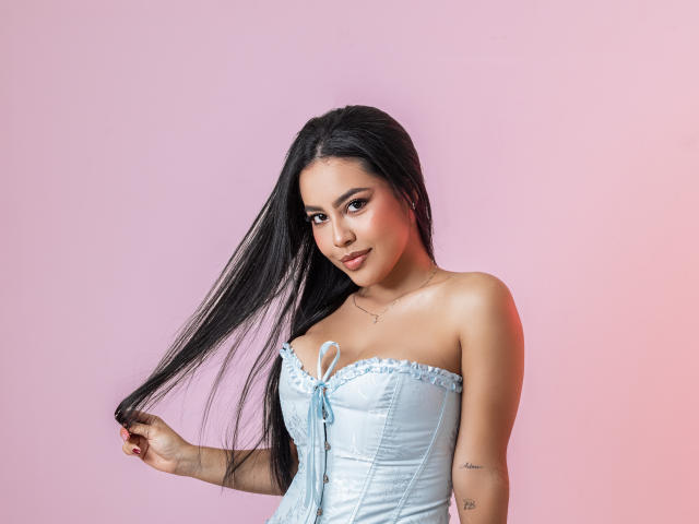 PaolaYKatty with long black hair, wearing a light blue strapless corset, holding her hair, showing a tattoo on her left arm, and smiling subtly. Her tits are small.