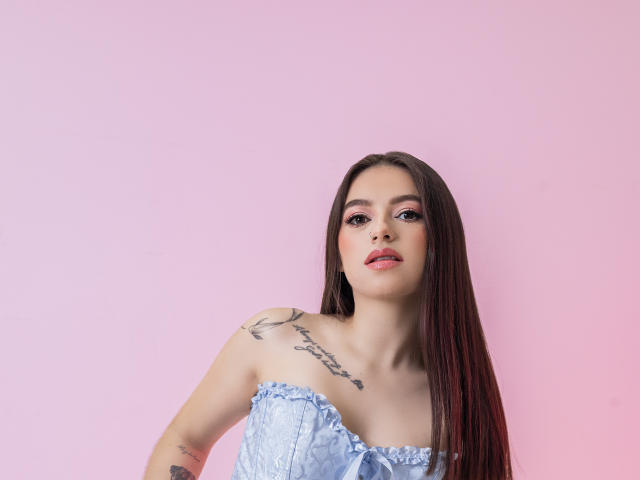 PaolaYKatty, avec de longs cheveux bruns, un maquillage rose clair, des tatouages sur les épaules et le bras, portant un haut bleu sans bretelles, avec un fond rose, regardant la caméra avec la bouche légèrement ouverte..