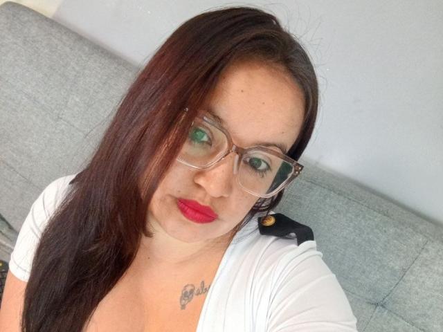 GinebraVargas with red lipstick, brown hair, glasses, white top, and a chest tattoo, looking at the camera with a slight smile, sitting on a gray couch.