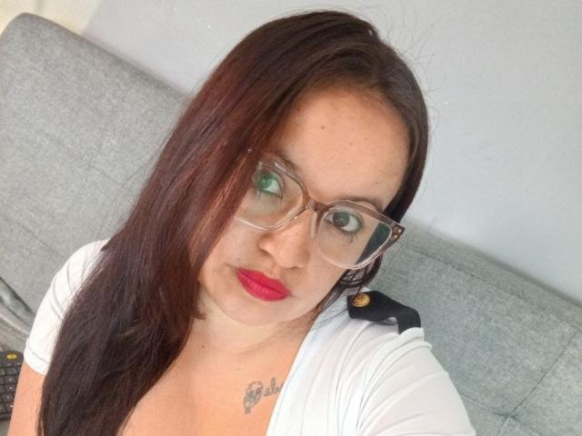 GinebraVargas has long brown hair, green eyes, and red lipstick. She's wearing glasses and a white top, with a small tattoo above her cleavage. She's sitting on a grey sofa.