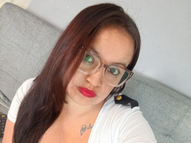 GinebraVargas has long brown hair, green eyes, red lipstick, clear glasses, white top, and a small tattoo on her chest. She's sitting on a gray couch.