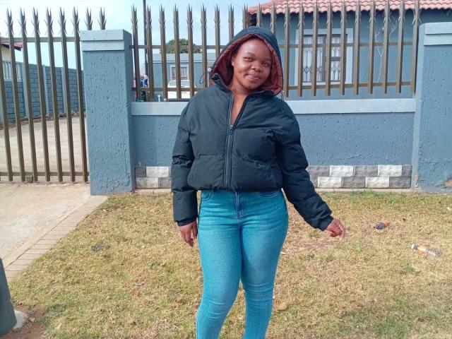 MandyRonny trägt eine dunkelgrüne, gepolsterte Jacke, eine rote Kapuze, blaue Jeans und steht auf Gras. Im Hintergrund ist ein Metallzaun zu sehen. Sie lächelt leicht, der rechte Arm ist herabgelassen, der linke Arm entspannt..