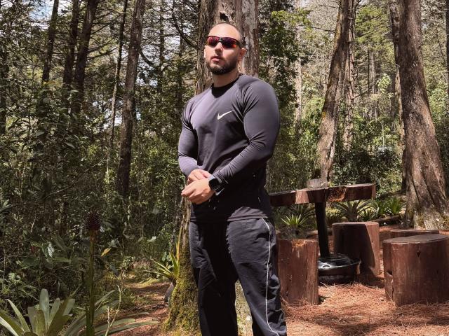 Frank Jones mit Bart, der ein schwarzes Langarmshirt, schwarze Hosen und rote Sonnenbrillen trägt, steht in einem Wald, die Hände gefaltet, eine Uhr am Handgelenk, und wirkt ernst..