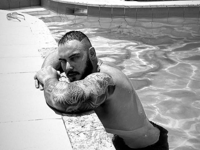 Frank Jones, mit Tattoos, lehnt sich am Poolrand ab, das Wasser reicht bis zur Taille, er wirkt ernst, hat einen kurzen Bart und nasses Haar. Es ist ein Schwarz-Weiß-Foto. Seine Arme liegen auf dem Poolrand, er trägt dunkle Badehosen..