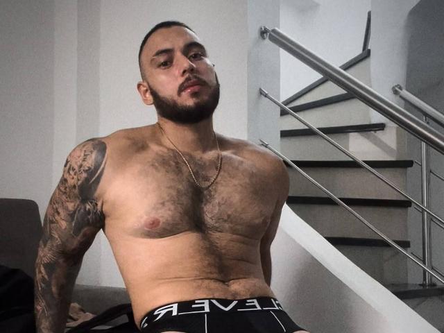 Frank Jones ist ein muskulöser Mann mit behaarter Brust, der Tattoos am rechten Arm hat, schwarze und weiße Unterwäsche trägt und auf einer Treppe mit einem Metallgeländer sitzt..