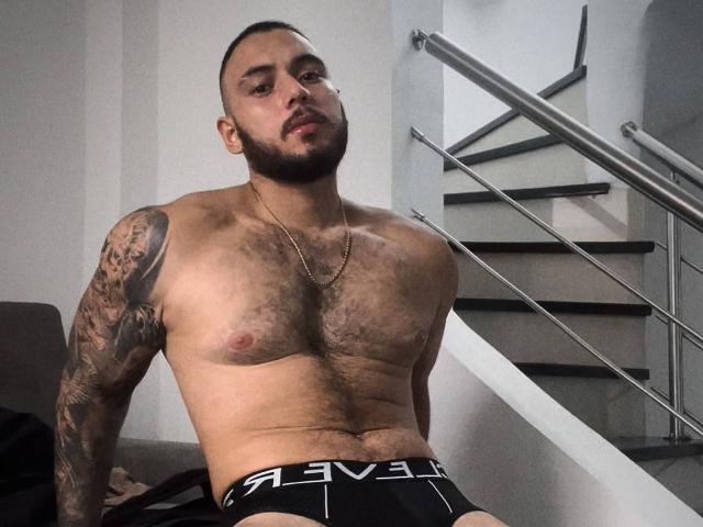 Frank Jones, senza camicia e con il petto peloso, un braccio tatuato e uno slip nero con l'elastico che mostra la lettera "P", è seduto su un divano, guardando la telecamera, con una scala sullo sfondo..