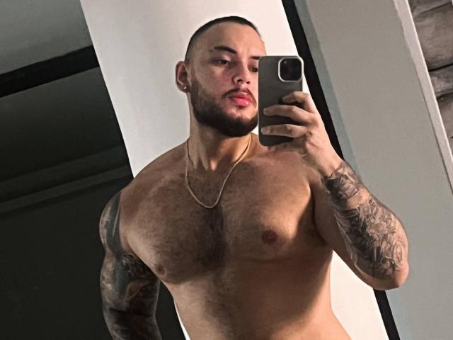 Frank Jones, ohne Hemd und mit Tattoos, hält ein Telefon, trägt eine goldene Kette, macht ein Selfie im Spiegel und zeigt eine behaarte Brust und einen gepflegten Bart, mit ernstem Gesichtsausdruck..