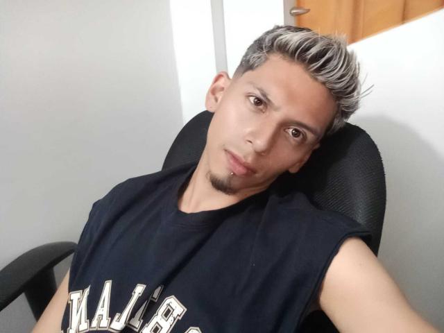 DereckMonroe, con i capelli grigi, un leggero pizzetto e un piercing al labbro, indossa una maglietta nera senza maniche, è seduto indietro su una sedia e guarda la telecamera..