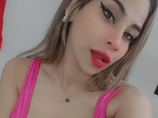 DayanaPrecious con rossetto rosso, eyeliner nero e una maglietta rosa, che mostra il seno, indossa una collana d'argento con un cuore viola e orecchini di diamanti, e guarda verso la fotocamera..